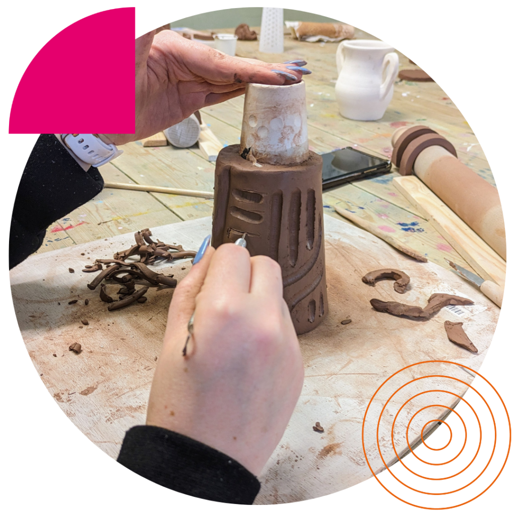In this photograph, we see hands engaged in decorating a ceramic piece. The object being worked on resembles a brown cylindrical tower with cut-out details, and the artist is applying what looks like white slip with a squeeze bottle, possibly for decorative or functional purposes such as attaching components. Shavings of clay are scattered on the table, which is also covered with brown paper. A rolled-up slab of clay, a smartphone, and a white ceramic mug are visible in the background, hinting at a multi-faceted pottery project in progress.