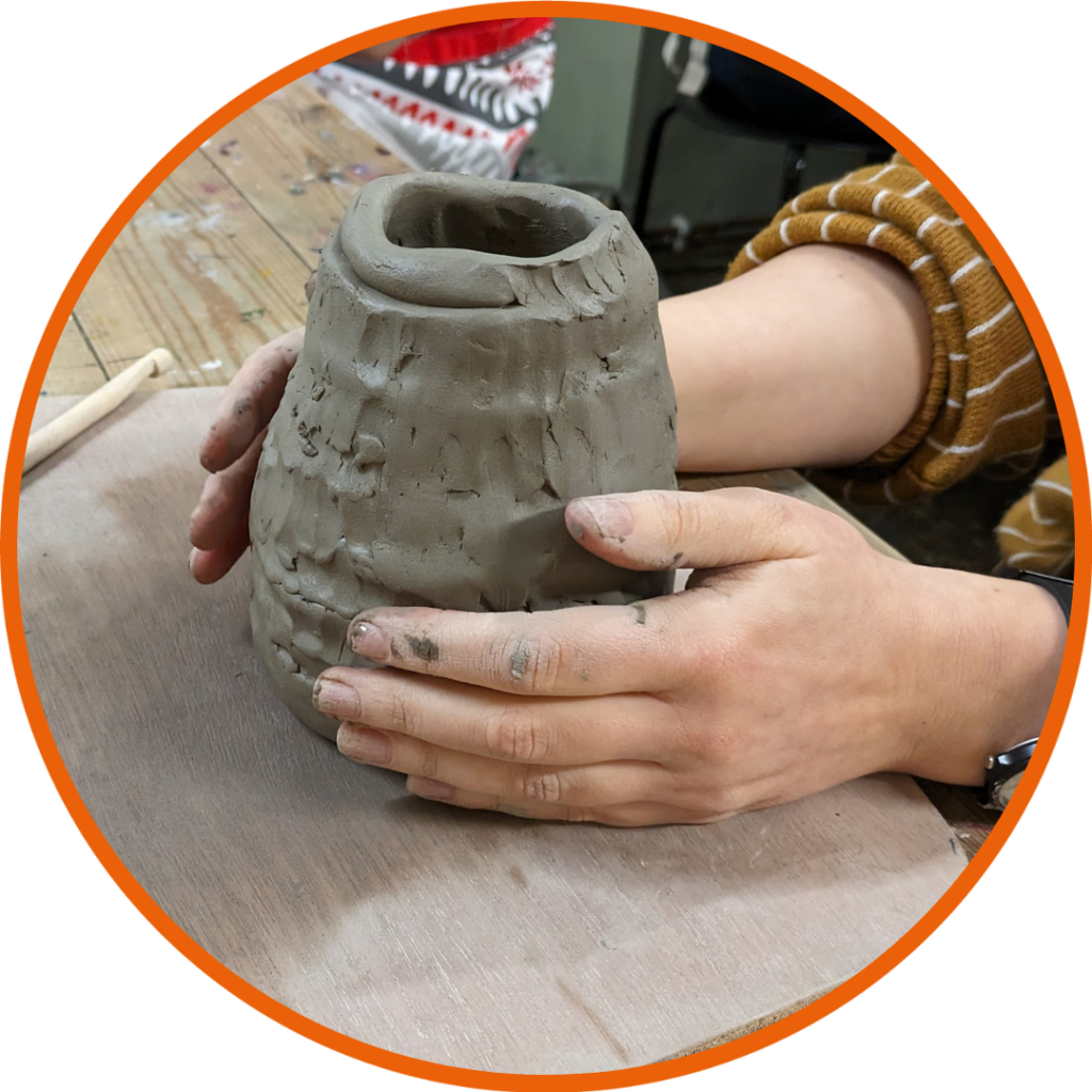 The focus here is on hands that are finishing the surface of a grey clay object. The hands gently touch the clay surface, which exhibits textural details resembling scallops or feathers. The object appears to be a hand-built pottery vase or pot with an organic, uneven form. It sits on a wooden table protected by brown paper, surrounded by various clay-working tools like a wooden rib and a metal kidney scraper. To the side, we see a rolled clay coil and a brown bracelet on the wrist of one hand, adding a personal touch to the scene.