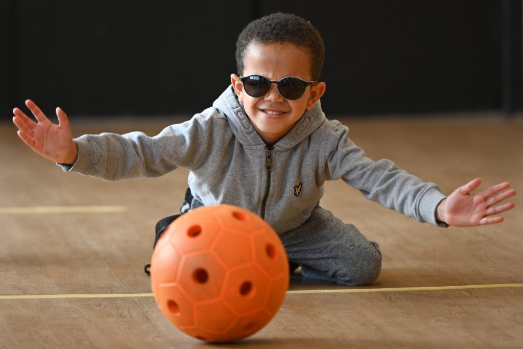 Image shows a young boy wearing dark glasses, kneeling on the floor as a ball rolls towards him.