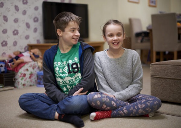 Image shows a boy and a girl sat cross-legged on the living room floor, both are smiling.