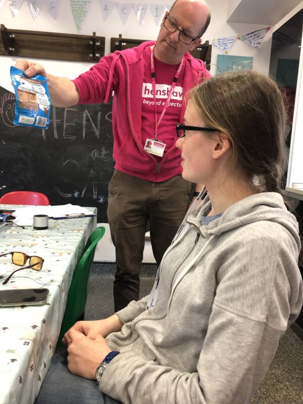 Images shows a young woman wearing some assitive tech to read some text being held by a man wearing a pink Henshaws t-shirt.
