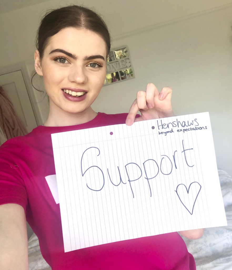 Image shows a young woman wearing a pink Henshaws tshirt, holding up a sign that says 'support.'