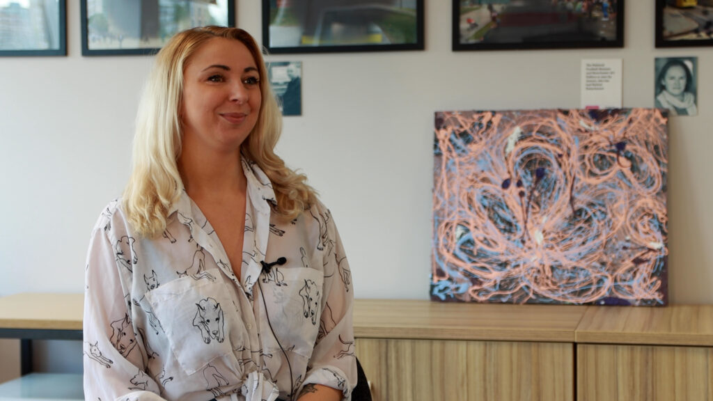 Mattie Screenshot from Insights smiling with artwork in background. The artwork is made from mixed media and in her Insights video she discusses what inspired her. The colours in the art are mixed light pastel colour swirls with dark blues and black in the background.