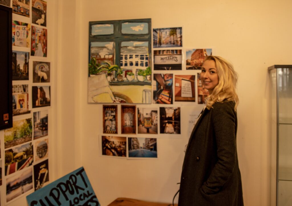 Matilda is stood smiling by her 'through a window' artwork at Afflecks Palace. The artwork displays what she can see through a window, including a radiator and chairs which are in front of the window.