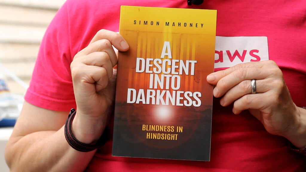 Markholding up a copy of Simon's book. The front cover reads "SIMON MAHONEY. A DESCENT INTO DARKNESS. BLINDNESS IN HINDSIGHT. The artwork shows the title of the book falling from the bright top of the book into darkness below