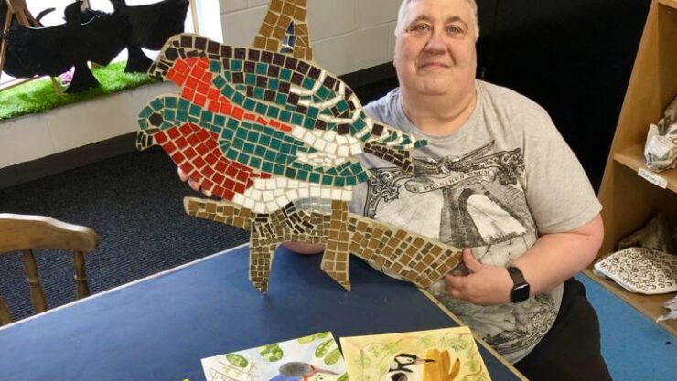 Anne is sat holding her two bird mosaic. In front of here are three watercolour pictures of birds and three Lino prints of the same birds in blues and greens